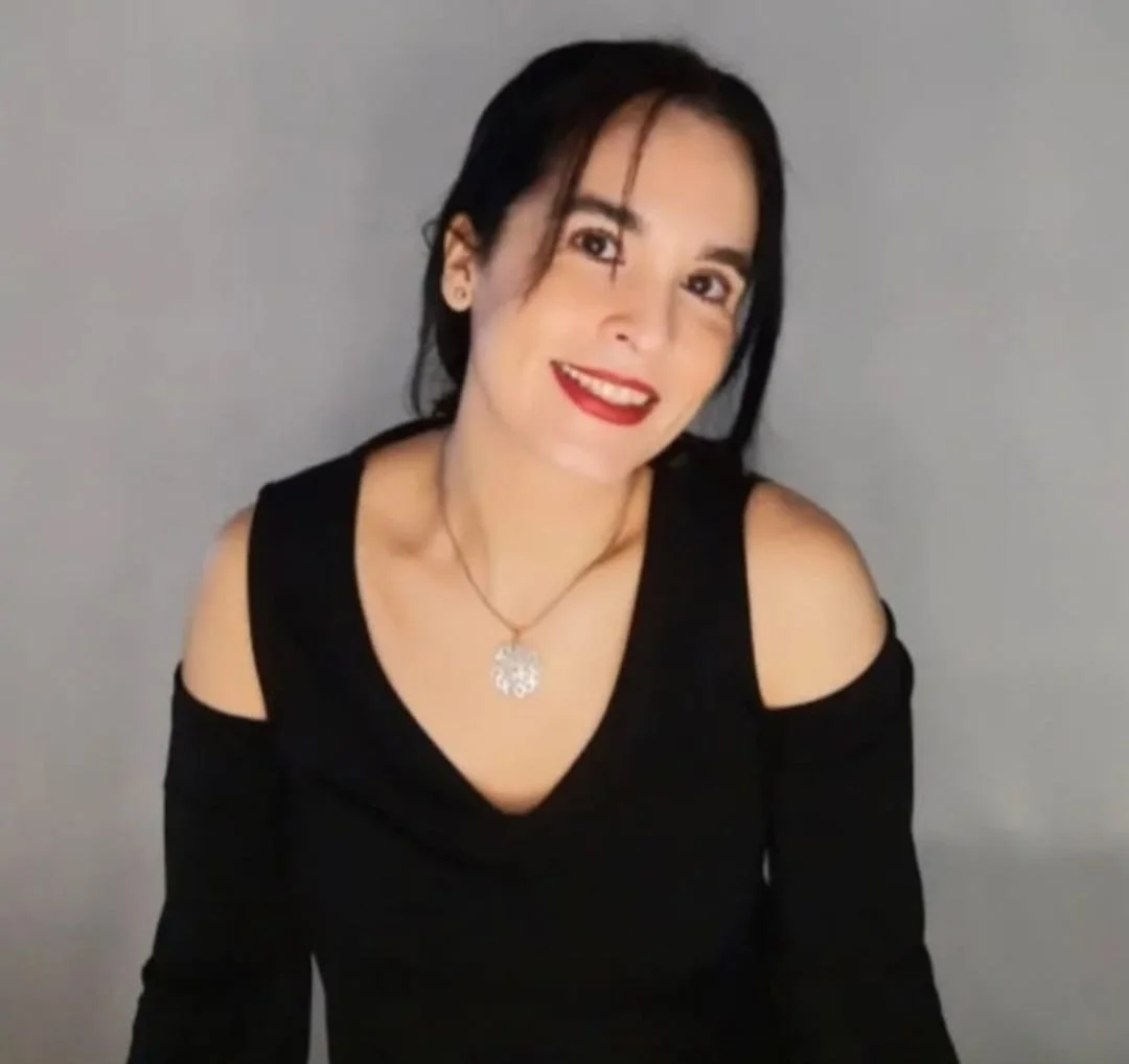 Mujer sonriente con cabello oscuro, vestido negro con hombros descubiertos, contra un fondo gris.