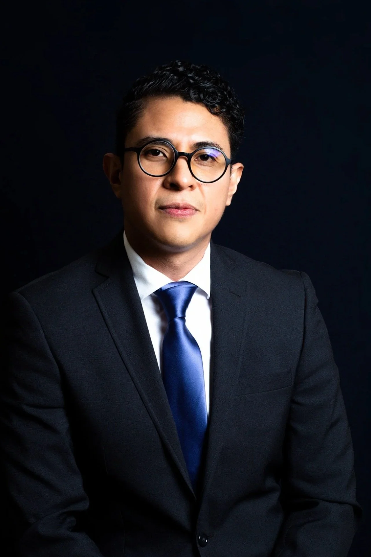 Hombre joven con cabello rizado y gafas redondas, vestido con saco oscuro, camisa blanca y corbata azul, posa contra fondo negro.