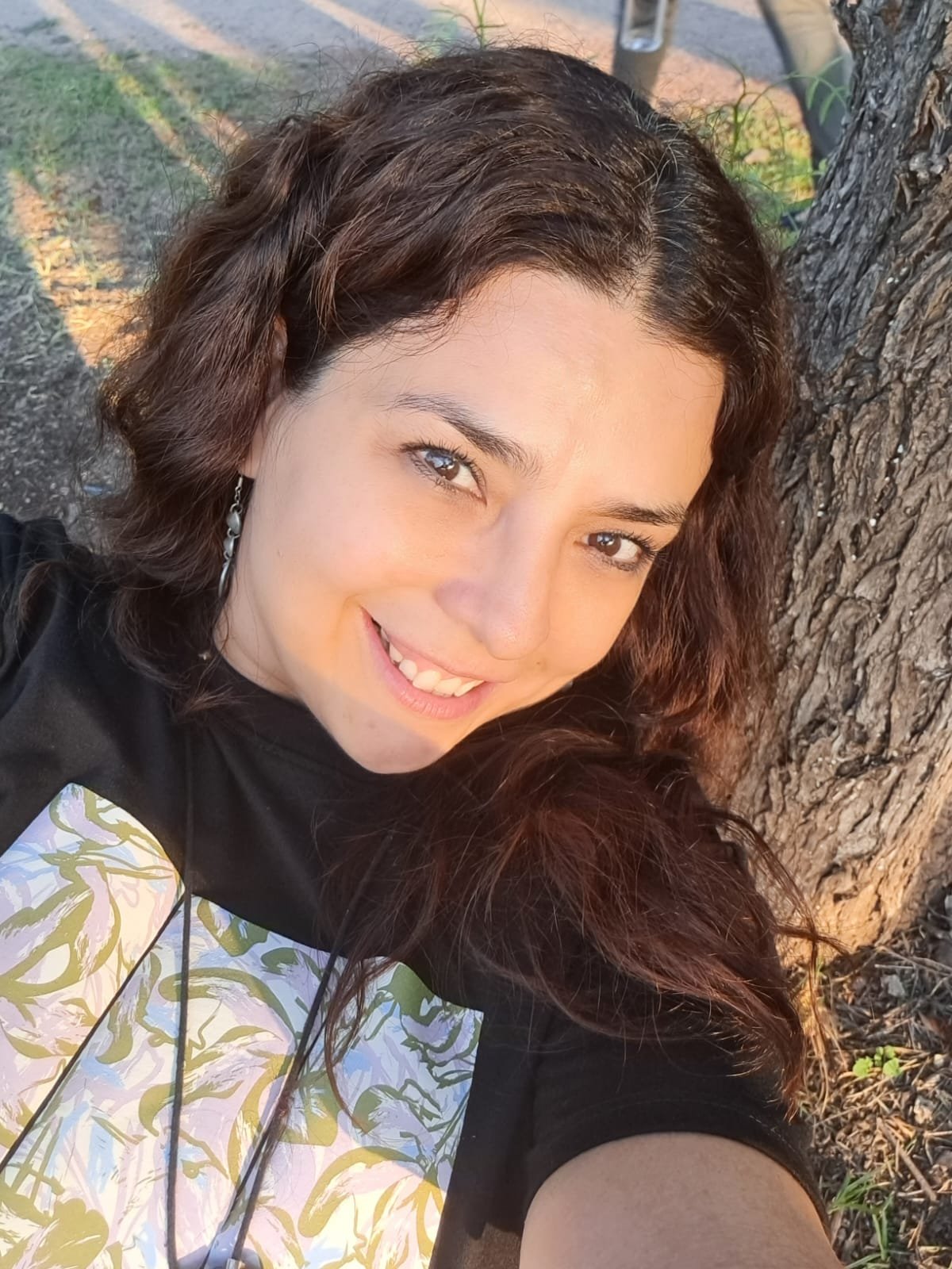 Retrato de una mujer sonriendo, con cabello rizado y oscuro, con fondo de árbol y césped, tomado en exterior durante el día.