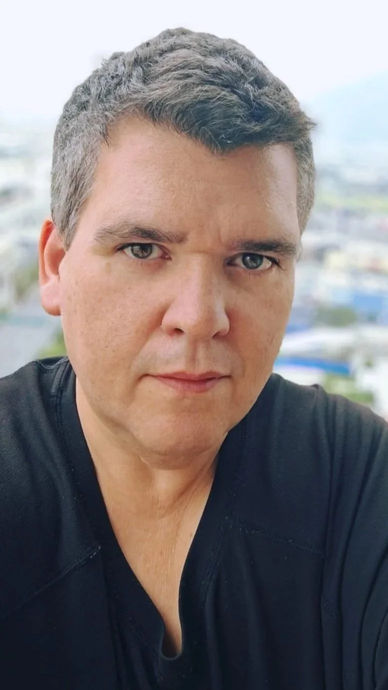 Retrato de un hombre con cabello castaño y canoso, piel clara, ojos claros, con expresión seria, vistiendo una camiseta negra, con fondo borroso de ciudad.